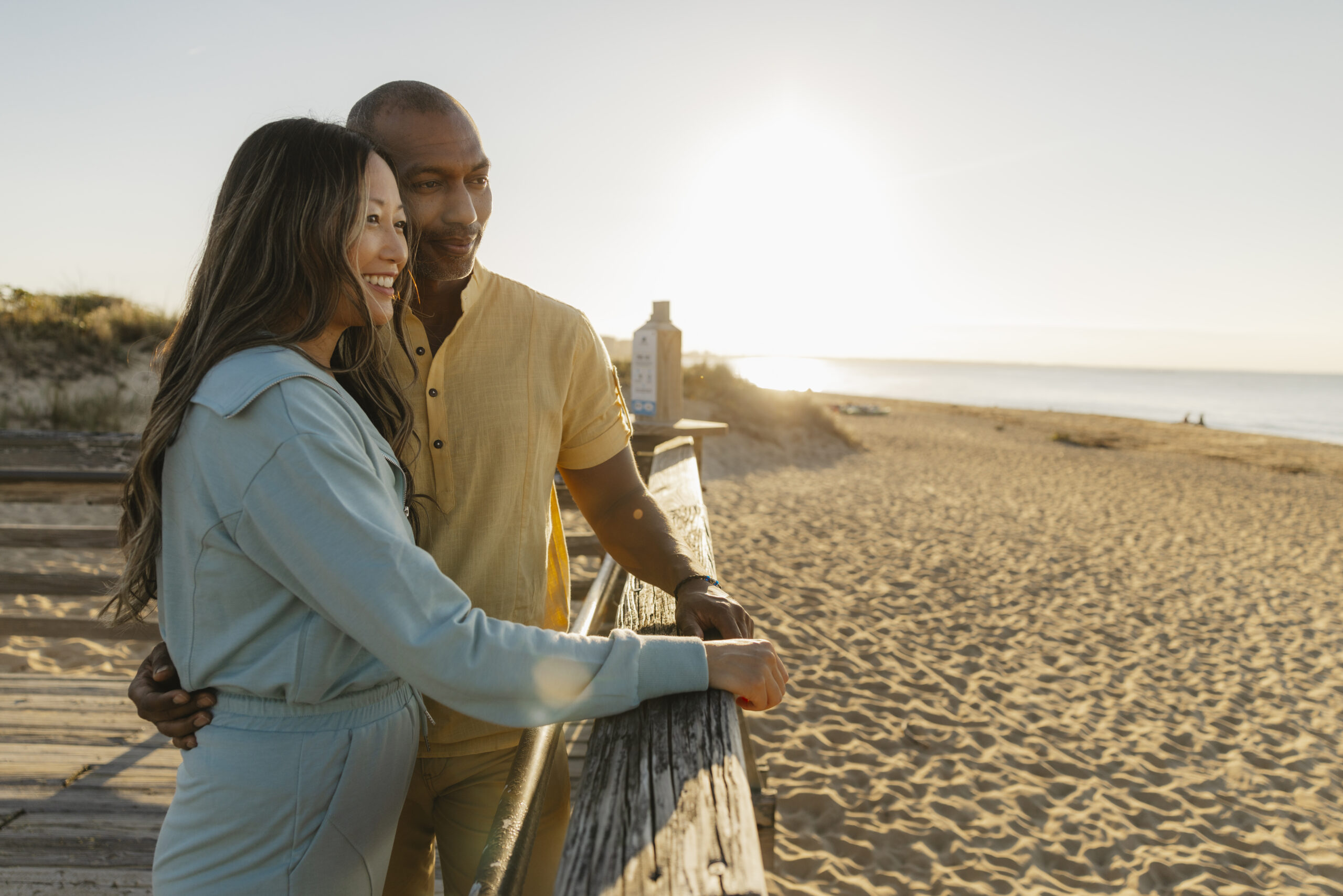 A couple enjoys a romantic moment on First Landing Beach's boardwalk at sunset, with soft sunlight illuminating the sandy shore.