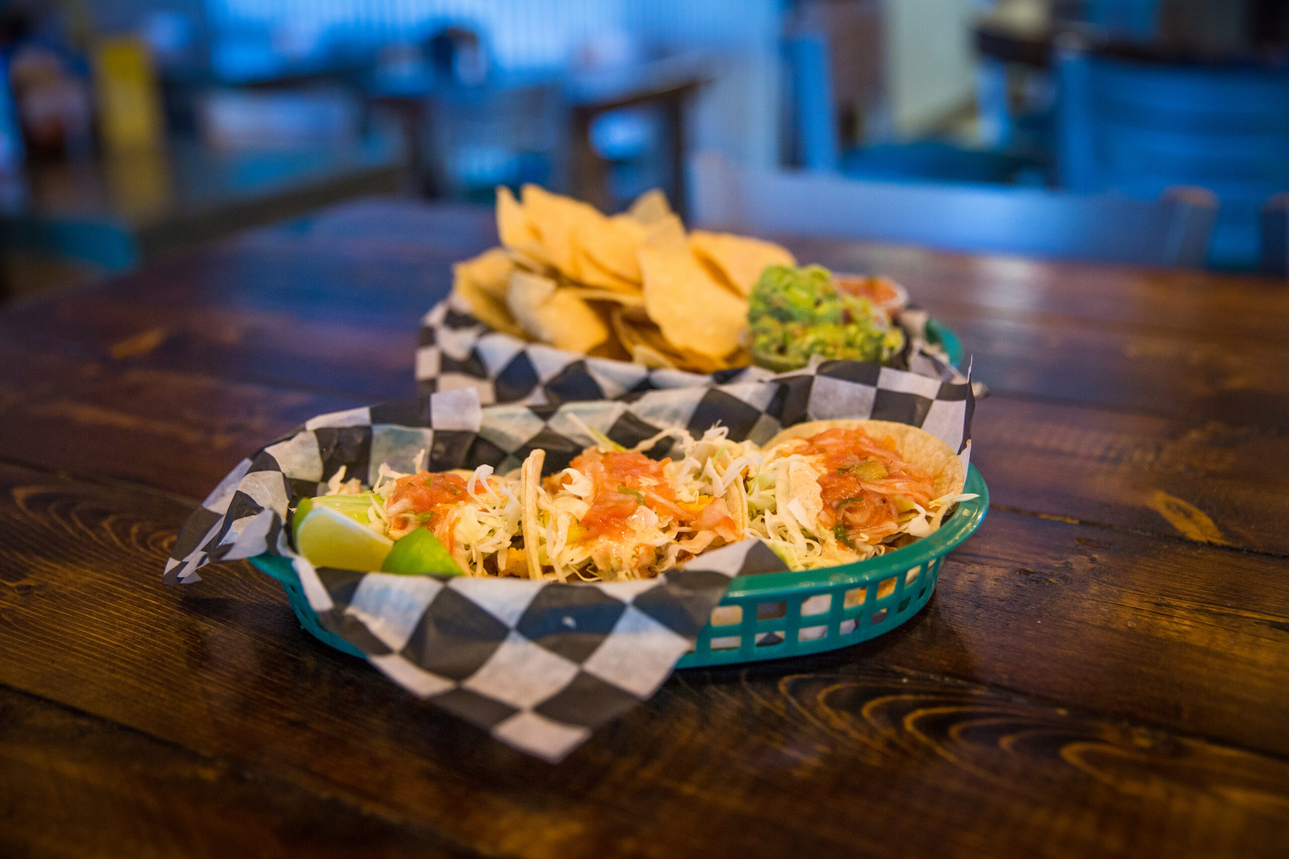 A basket with three tacos filled with lettuce, cheese, and tomatoes, and a side of lime on a wooden table. Another basket contains tortilla chips and guacamole.