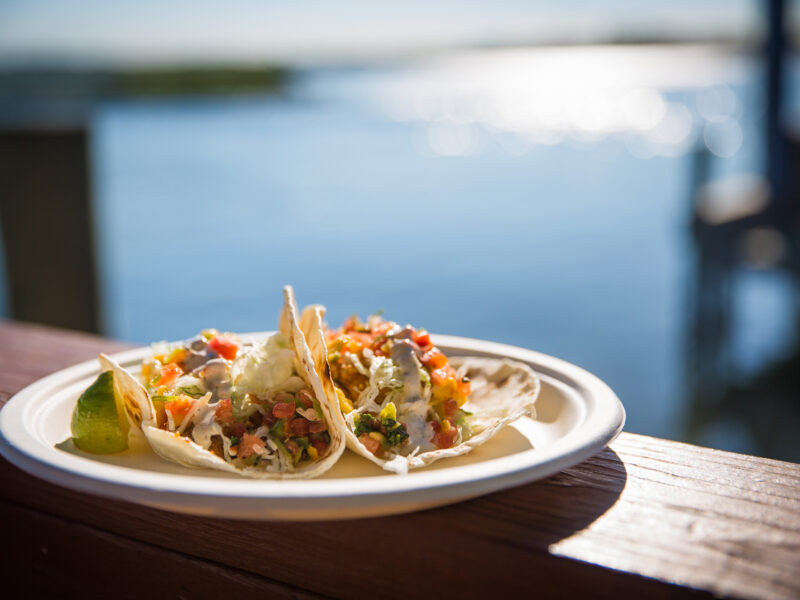Two tacos filled with vegetables and salsa on a plate, placed on a wooden railing by the water with a dock in the background.
