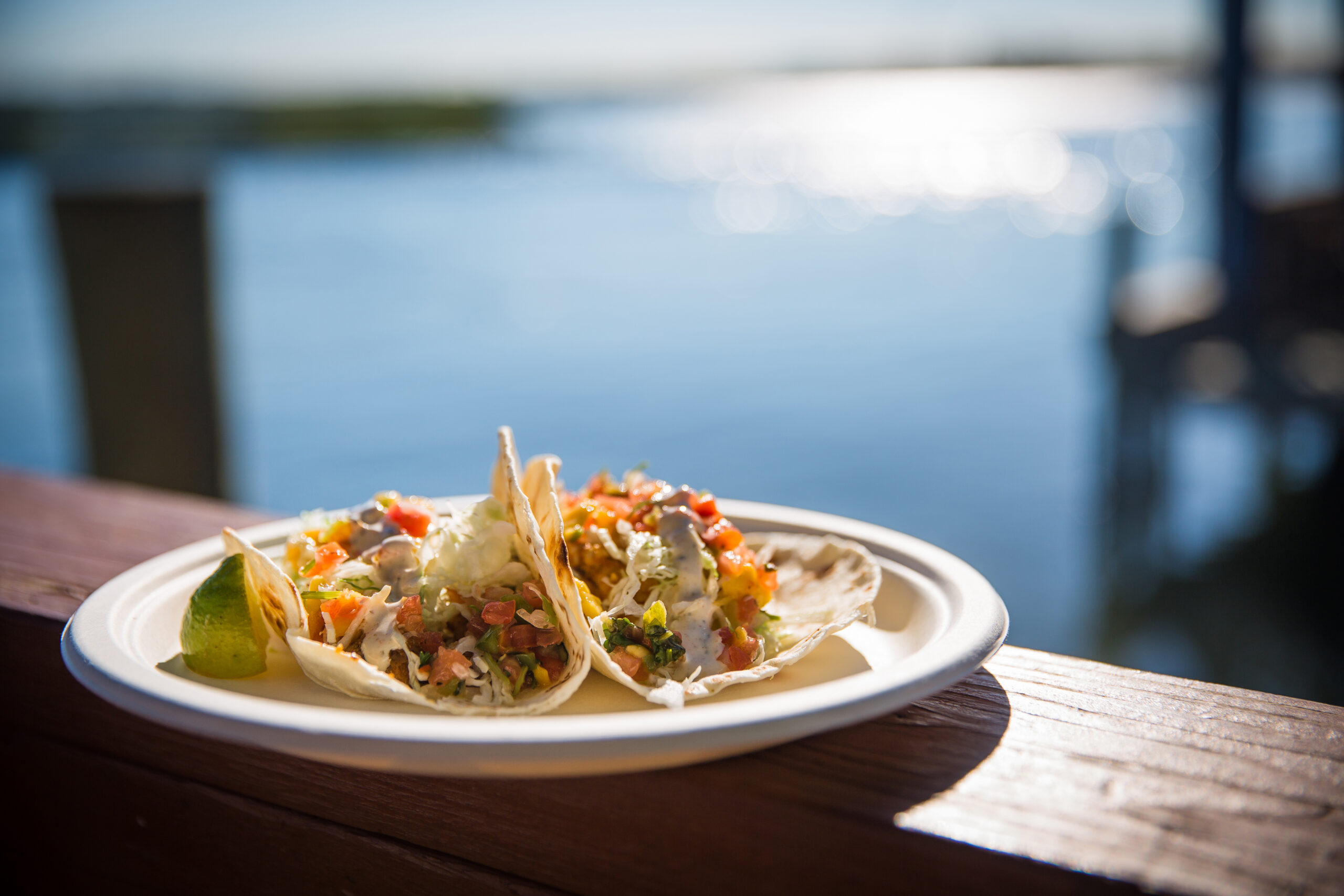 Two tacos filled with vegetables and salsa on a plate, placed on a wooden railing by the water with a dock in the background.