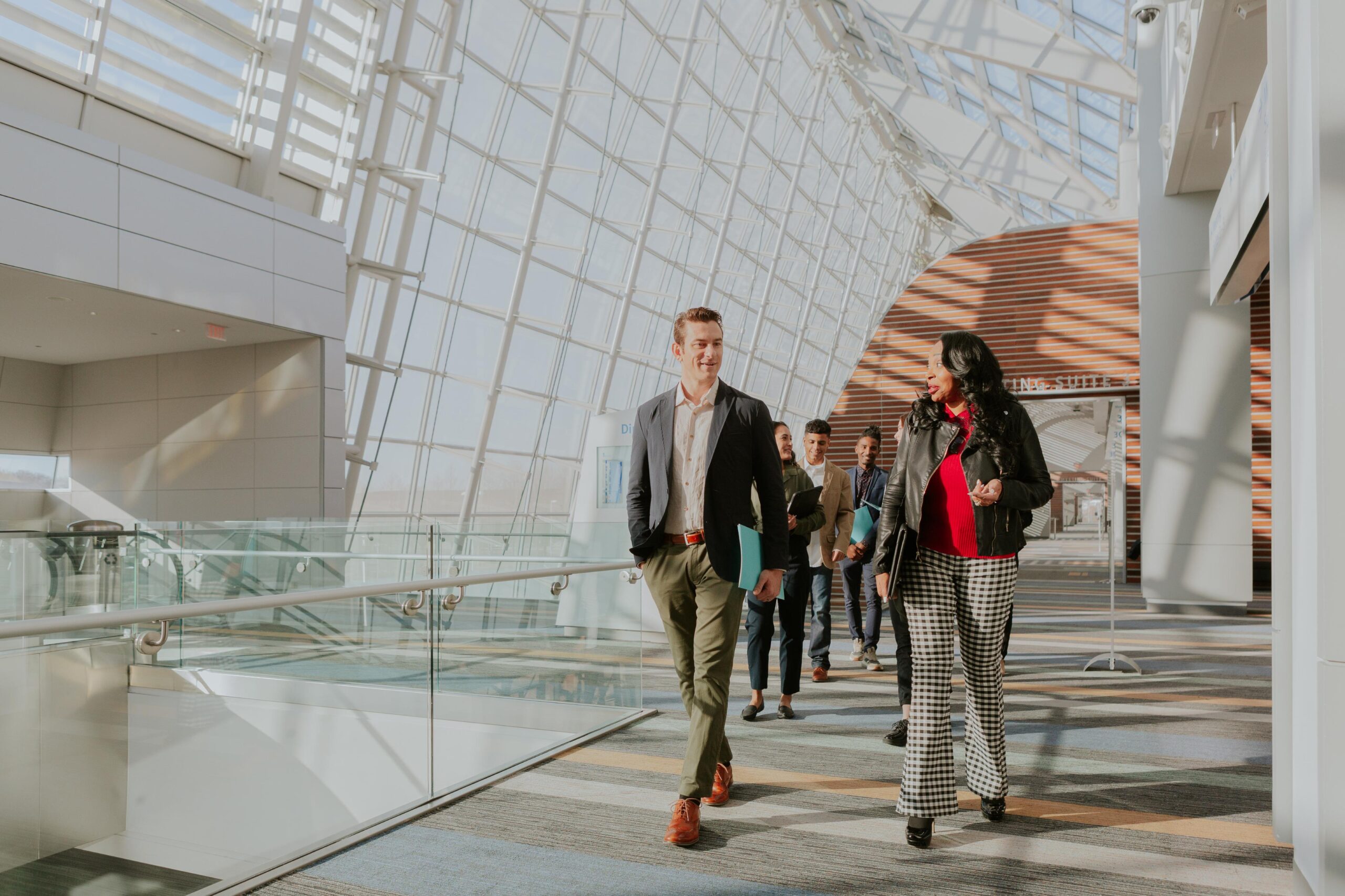 A group of professionally dressed people walk through a sunlit hallway in a modern office building with glass walls and high ceilings.