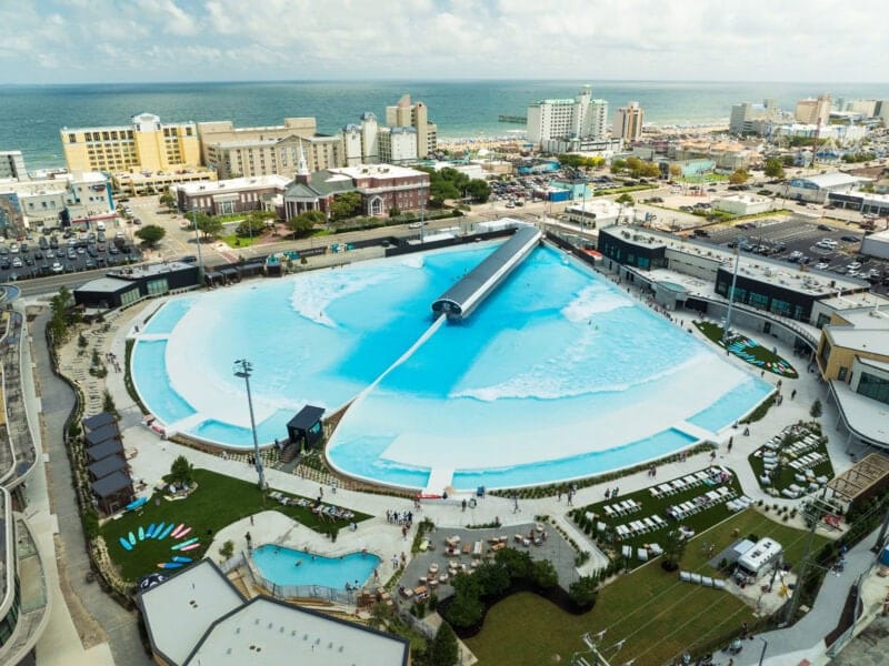 A large artificial wave pool with a central wave machine, surrounded by buildings, paths, and people, near a beach and cityscape.