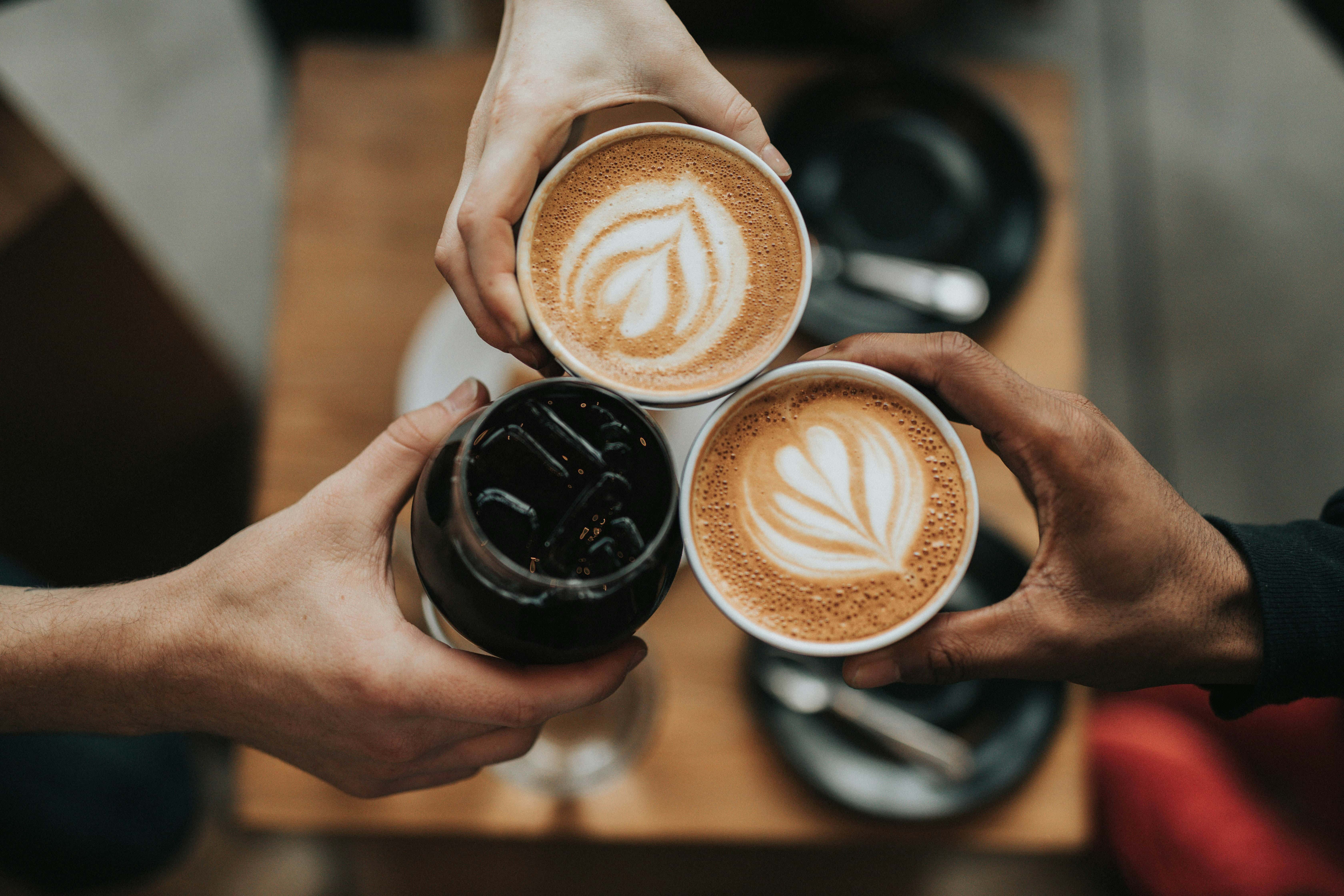 Three people holding drinks, two with lattes featuring latte art and one with a glass of iced coffee, over a wooden table with black saucers and plates.