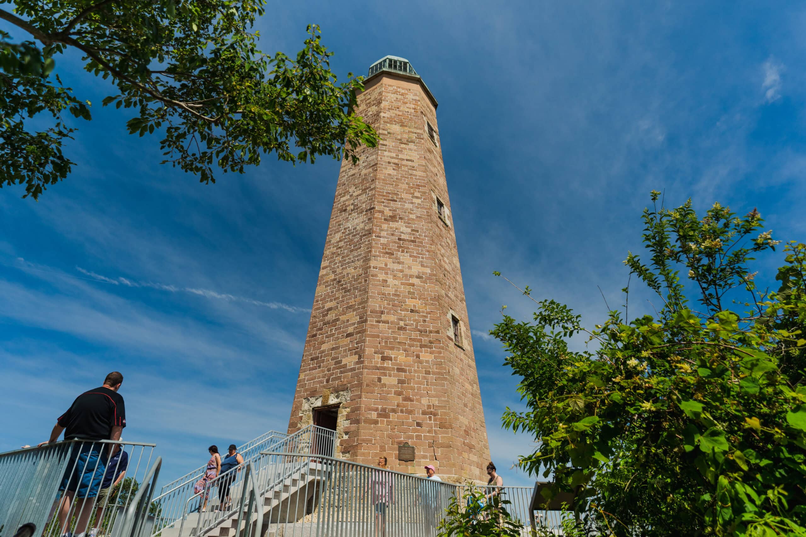 Visitors walk up stairs toward a tall, hexagonal stone lighthouse under a blue sky.