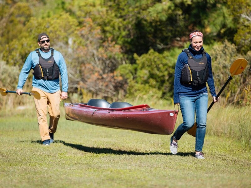 A man and a woman in life vests carry a tandem kayak across a grassy field while holding paddles.