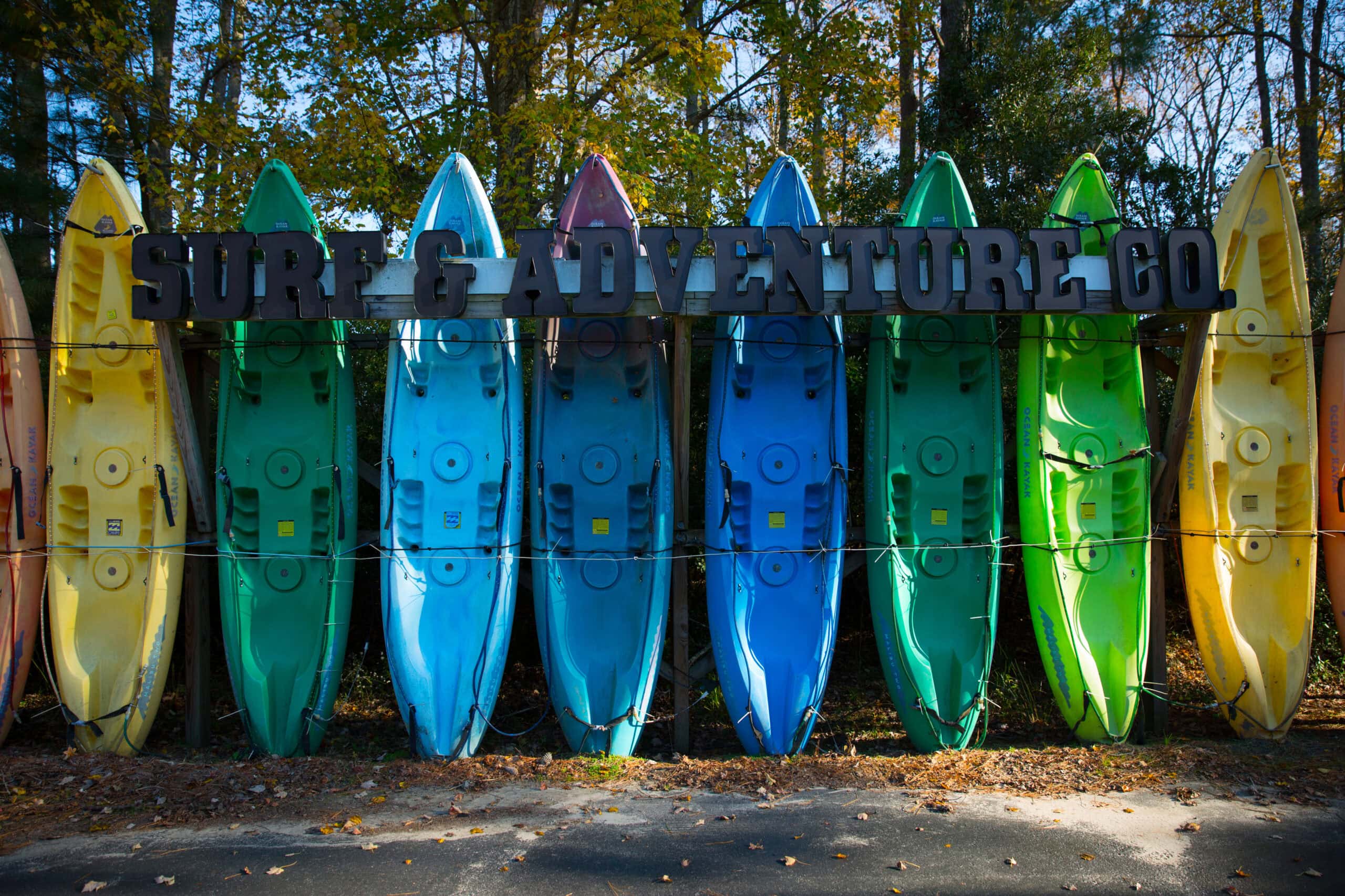 A rack of nine colorful kayaks hanging vertically under a sign that reads “SURF & ADVENTURE CO.”