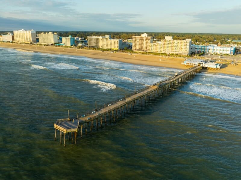 Aerial view of a long wooden pier extending into the ocean from a sandy beach lined with hotels.