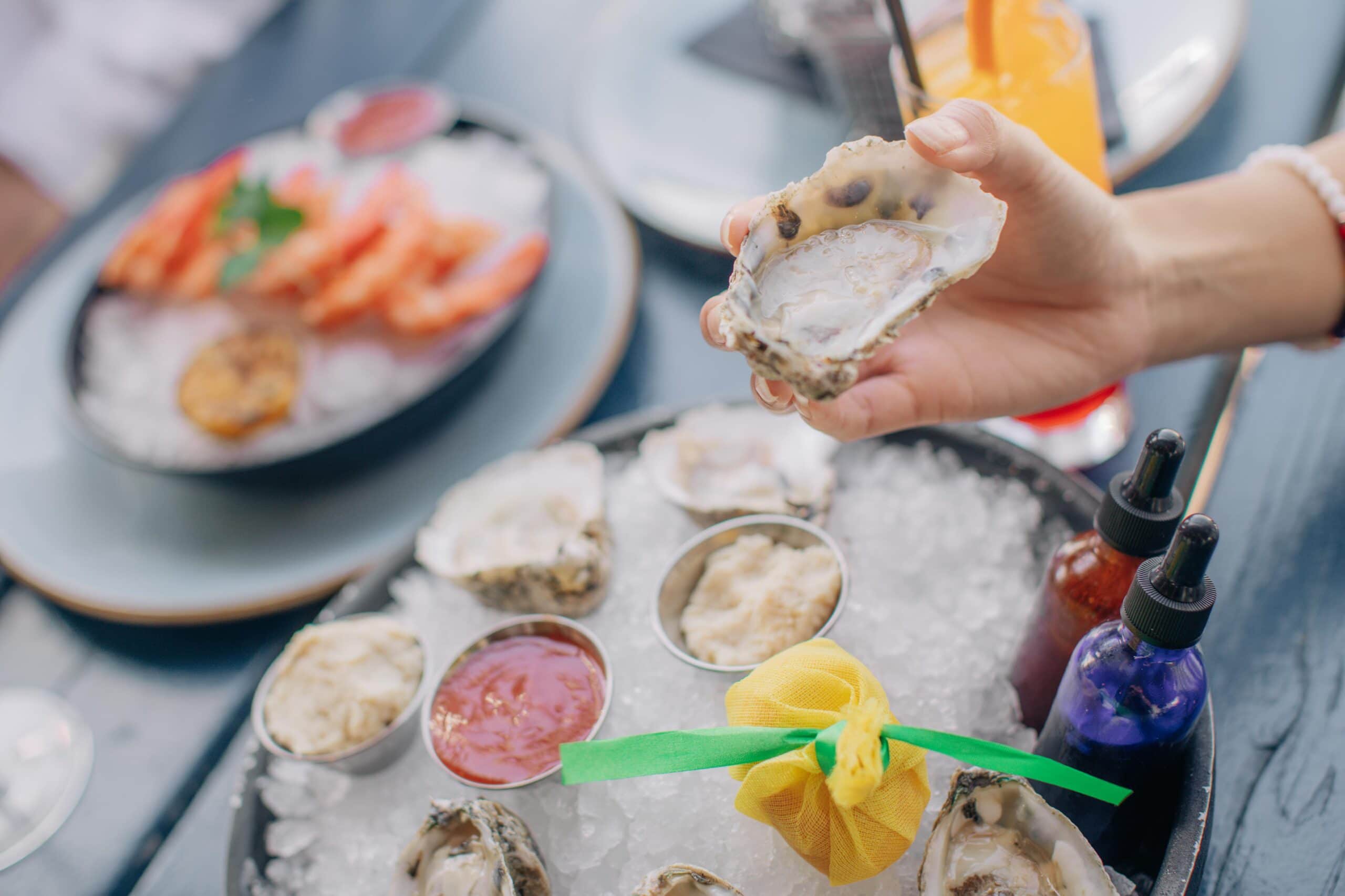 A hand holds an open oyster over a platter of raw oysters on ice, served with sauce and a lemon wrapped in yellow mesh.